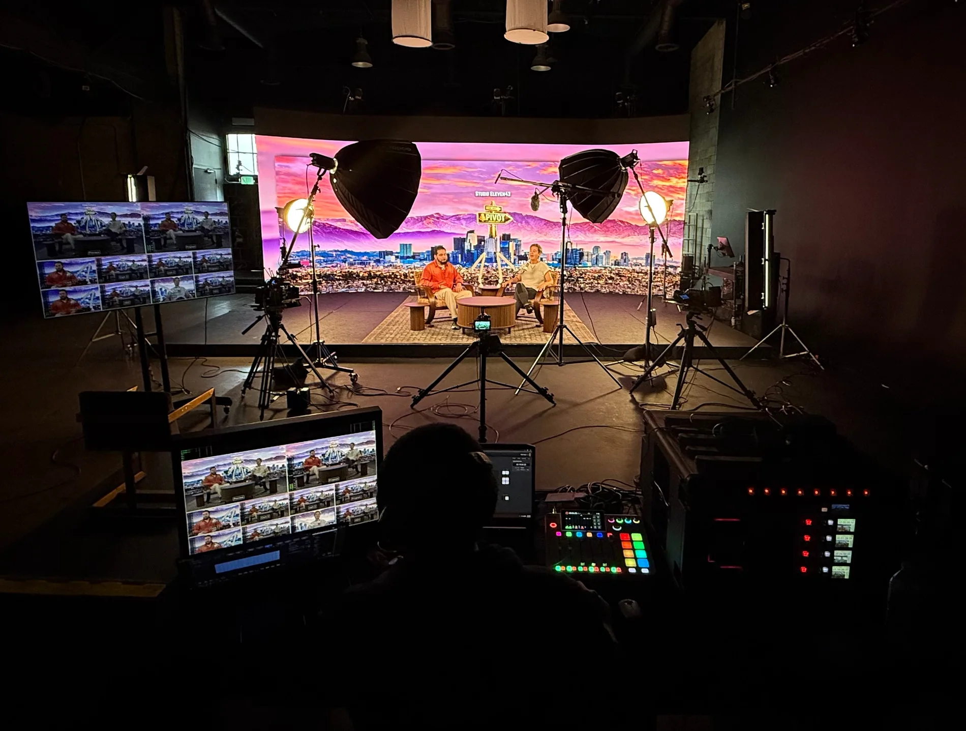 Podcast studio in Los Angeles: two hosts recording on the Hollywood LED volume stage at Podcast Pros LA, lit with softboxes, four Blackmagic 4K cameras on the floor and the live multiview, ATEM switcher and Rodecaster Pro audio console in the control room foreground.