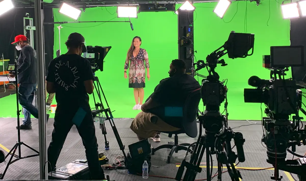 On location: talent on a green screen stage reading from a broadcast teleprompter, with crew and multiple cameras positioned around