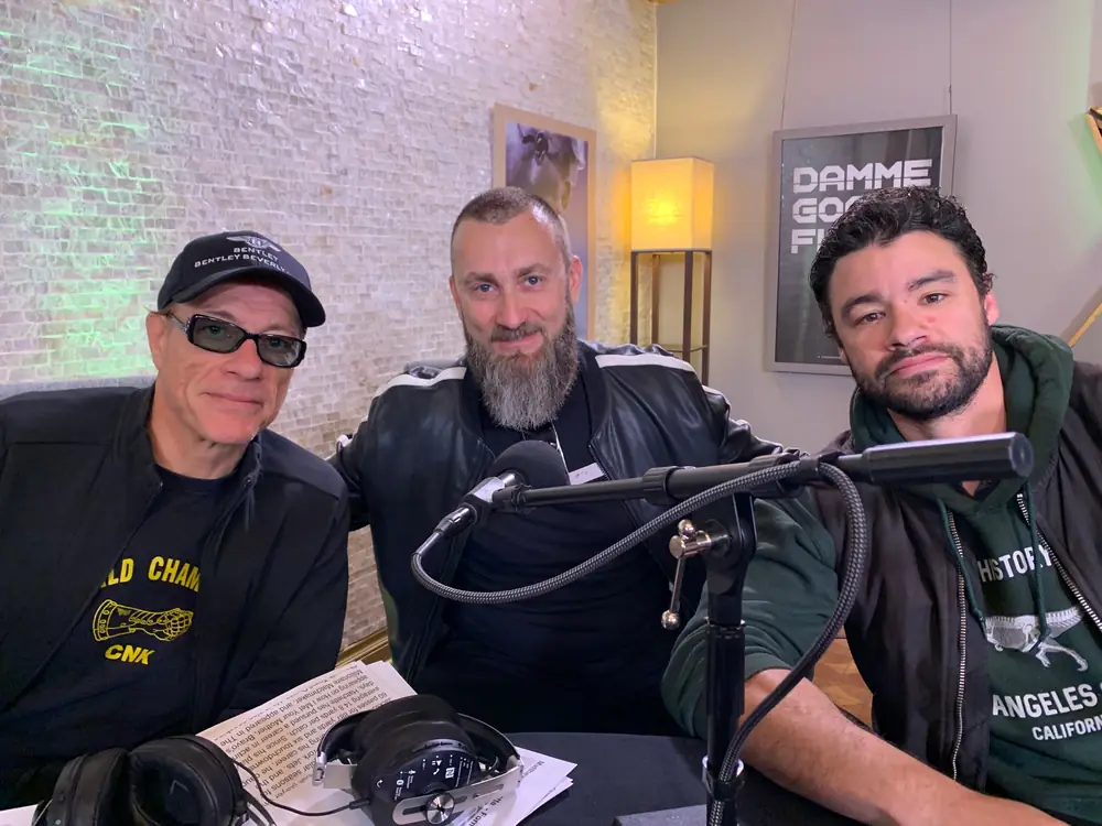 Three guests around a podcast table with broadcast microphones on boom arms in a dressed interview room